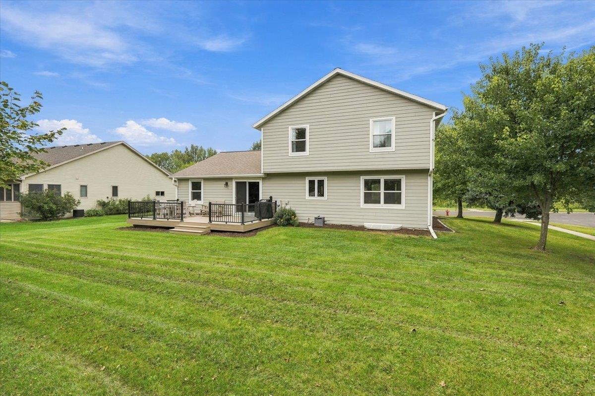 Rear of a two-story home with a raised deck, broad lawn, and mature trees.
