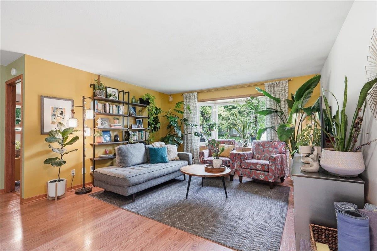 Living room with a gray sofa, patterned armchairs, and houseplants by a large window.