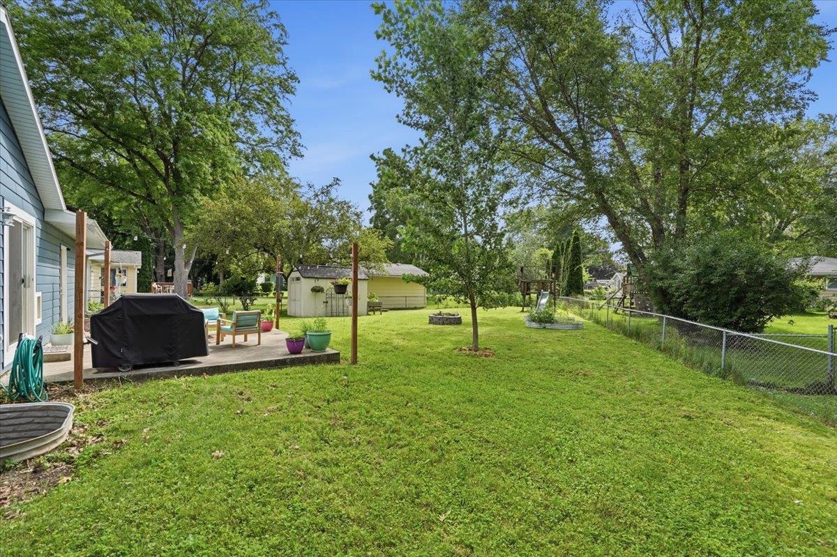 Grassy backyard with a patio, storage shed, mature trees, and children's playset.
