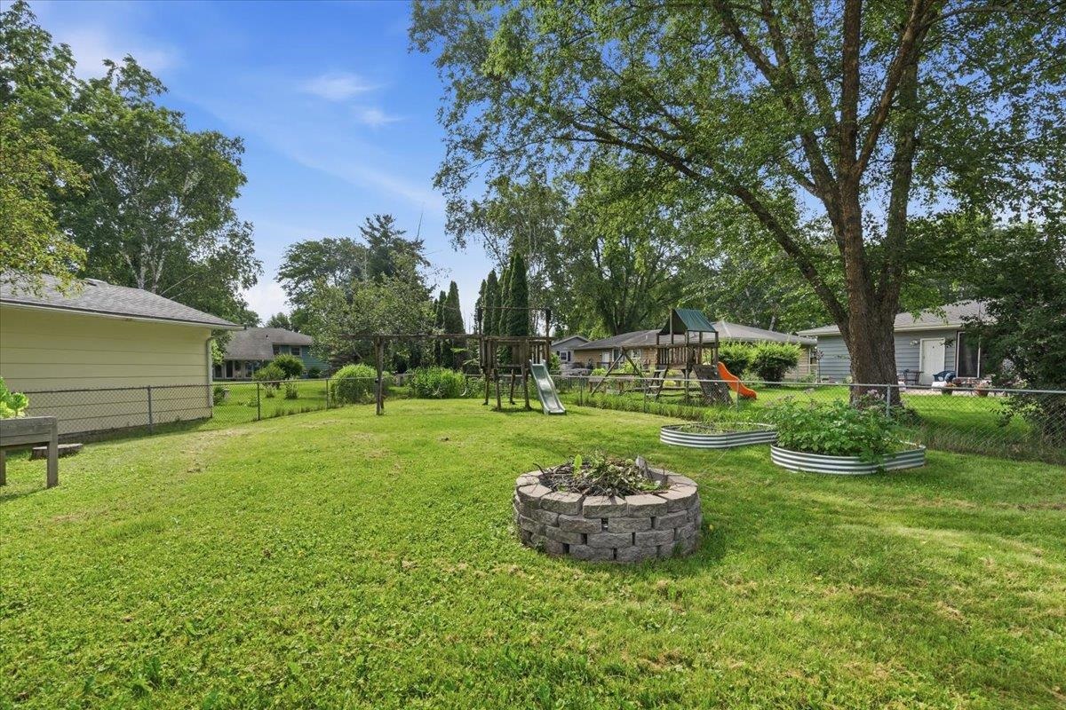 Backyard with a wooden playset, raised garden beds, and a stone fire pit beneath mature trees.