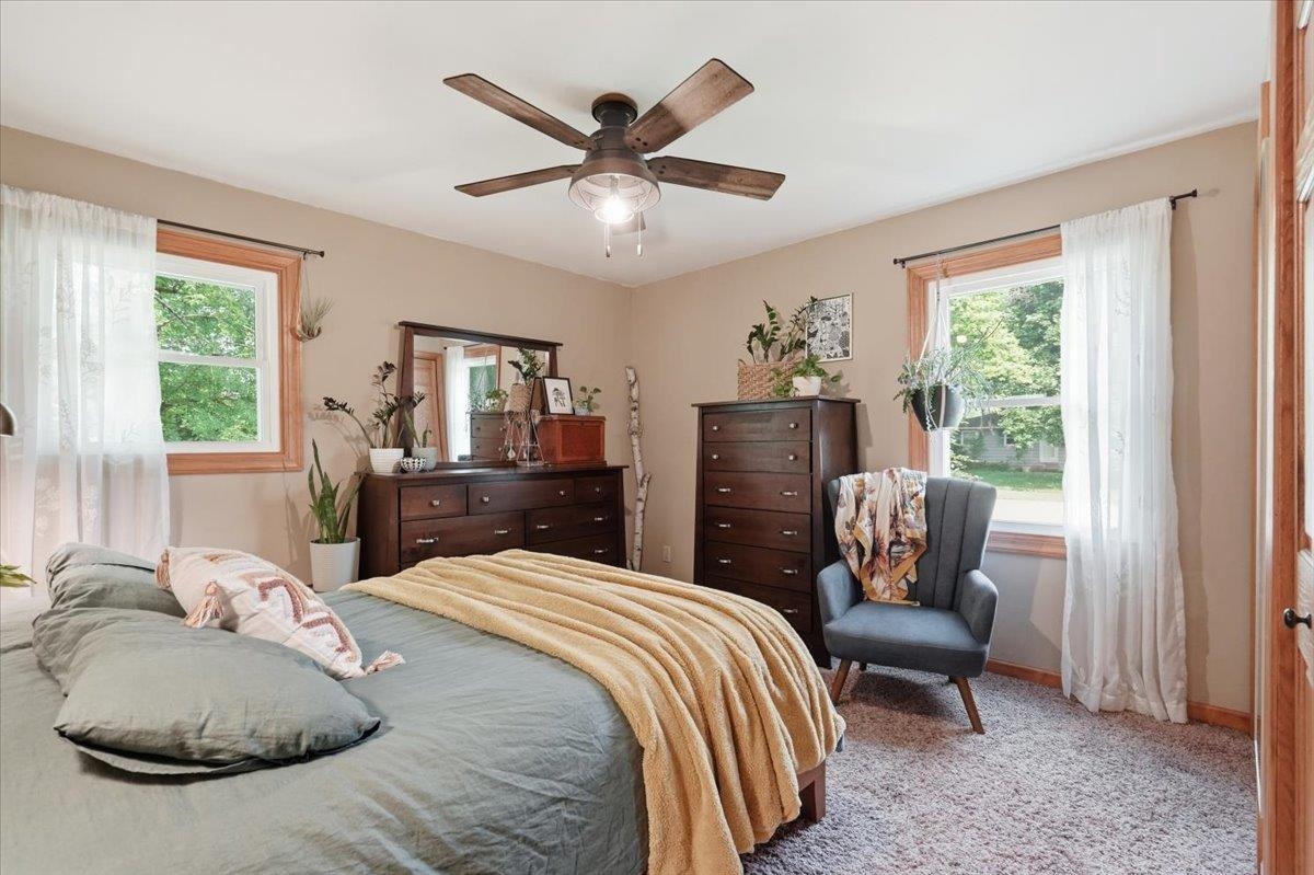 Bedroom with sage bedding, dark wood dressers, and a gray armchair between two curtained windows.