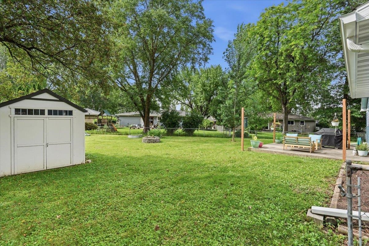 Grassy backyard with mature trees, a white shed, stone fire pit, and furnished patio.