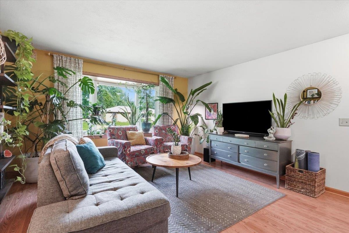 Living room with a gray sofa, floral armchairs, and leafy plants beside a large window.