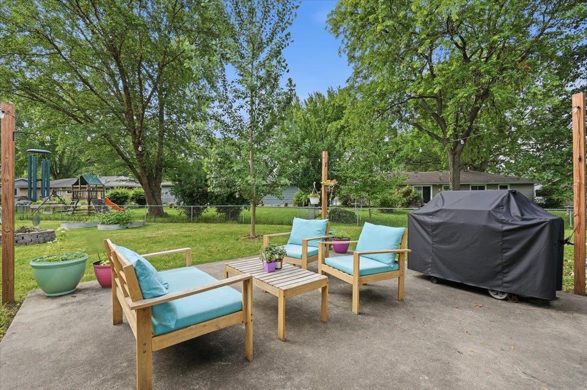 Backyard patio with turquoise-cushioned chairs, a covered grill, and mature trees.