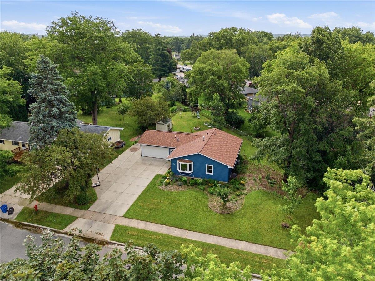 Aerial view of a blue home with an attached garage, wide driveway, and mature trees.