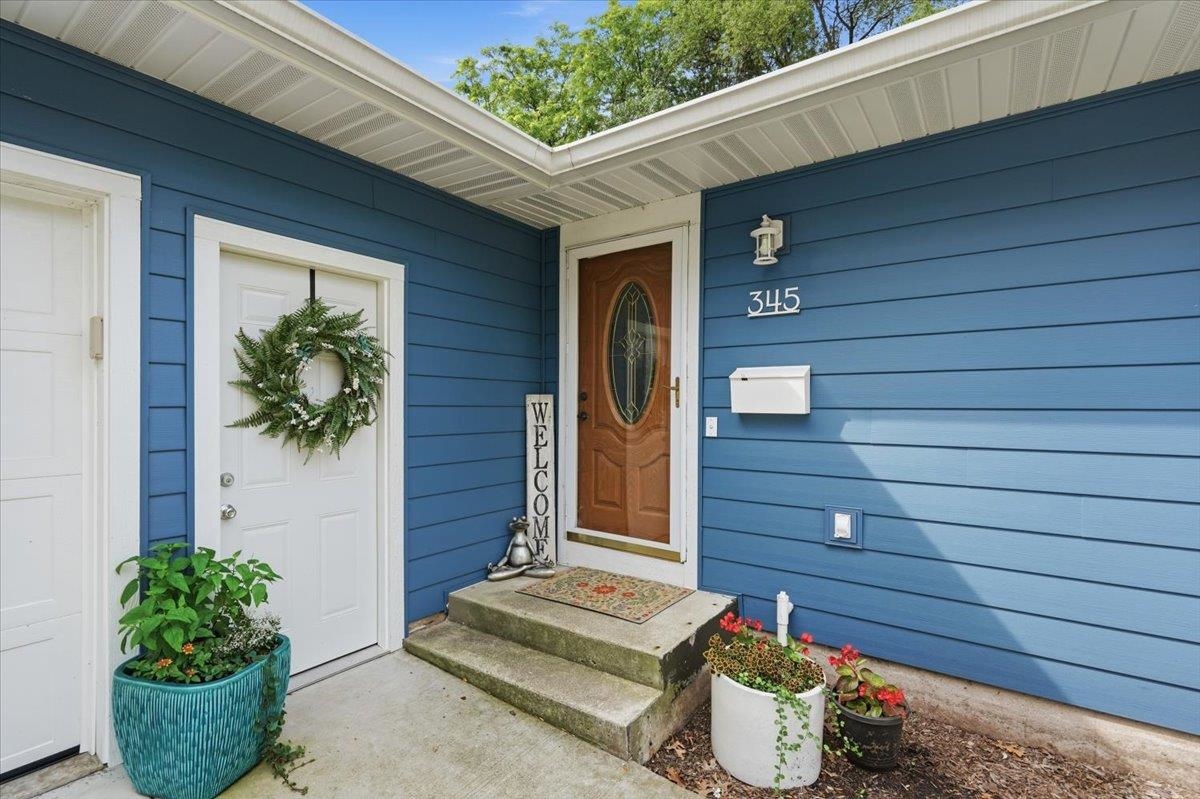 Blue-sided home entry with a brown oval-glass door, concrete steps, and potted flowers.