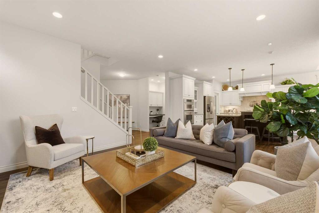 Open-concept living room with a gray sofa, cream armchairs, and a staircase beside the kitchen.