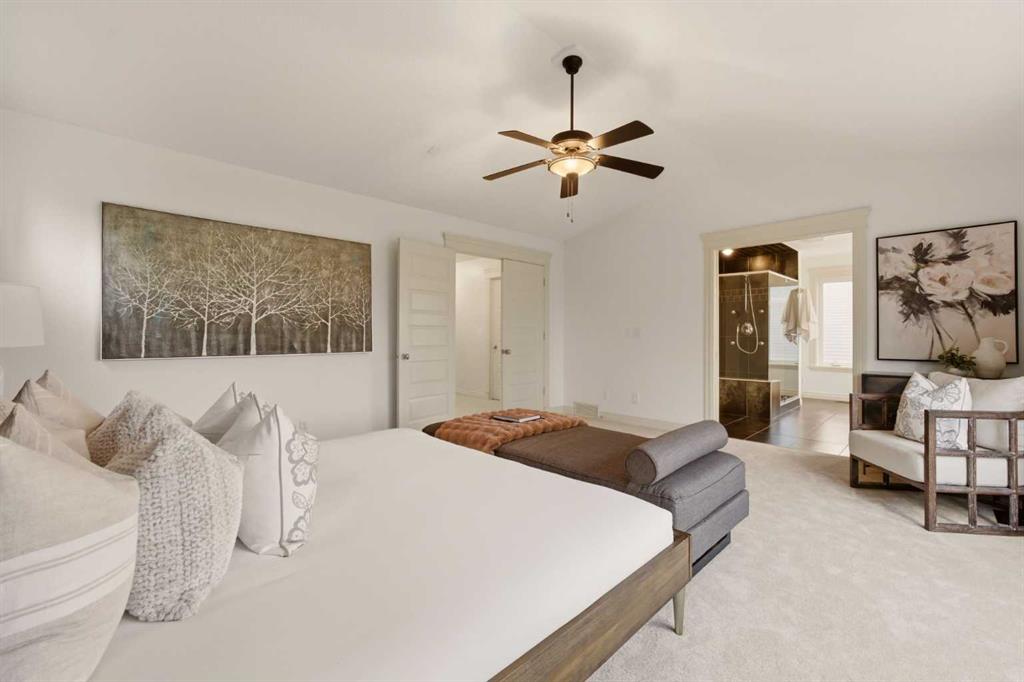 Staged bedroom with a large white bed, gray bench, ceiling fan, and ensuite bathroom doorway.