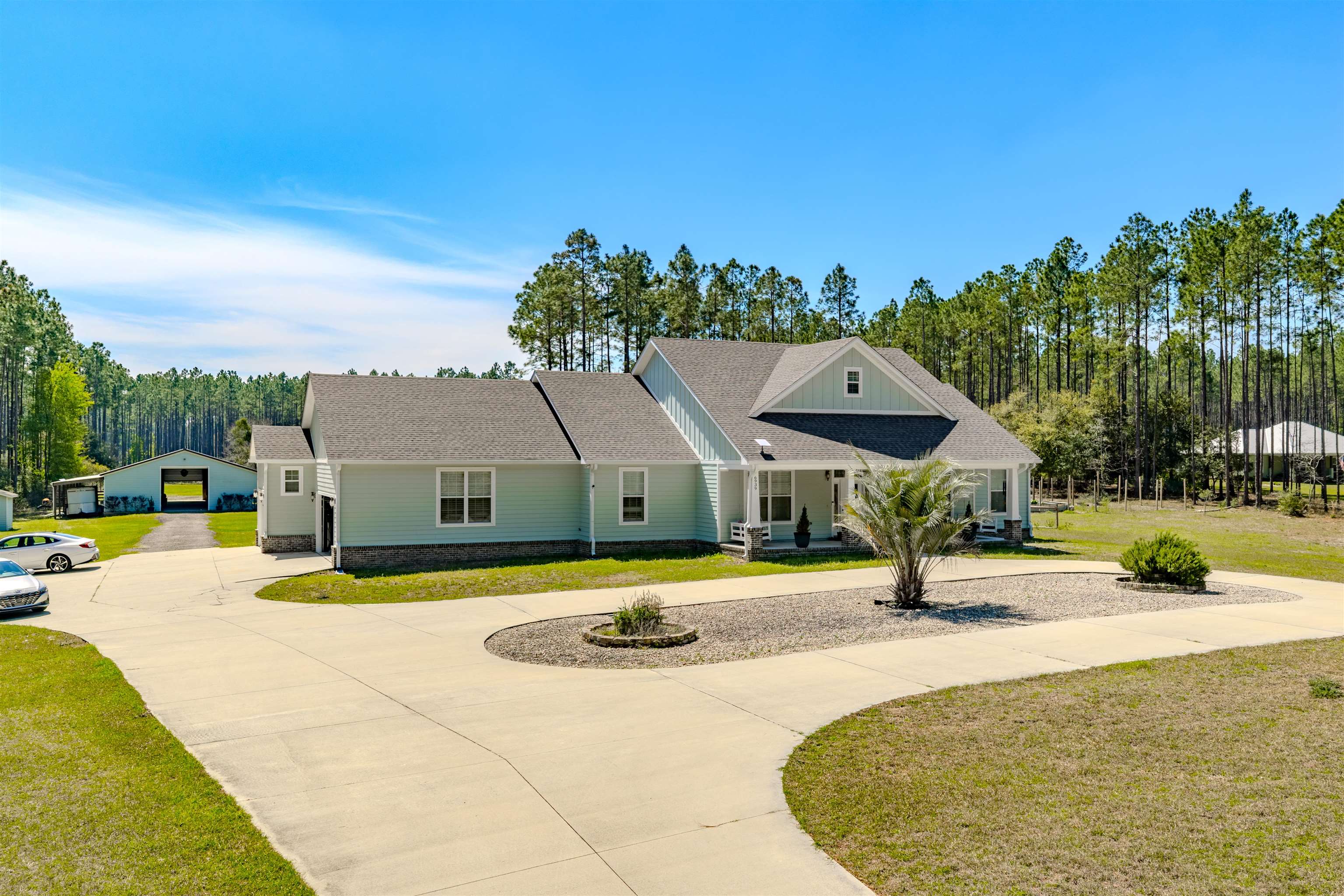Pale blue house with a covered porch, curved driveway, gravel landscaping, and pine trees behind it.