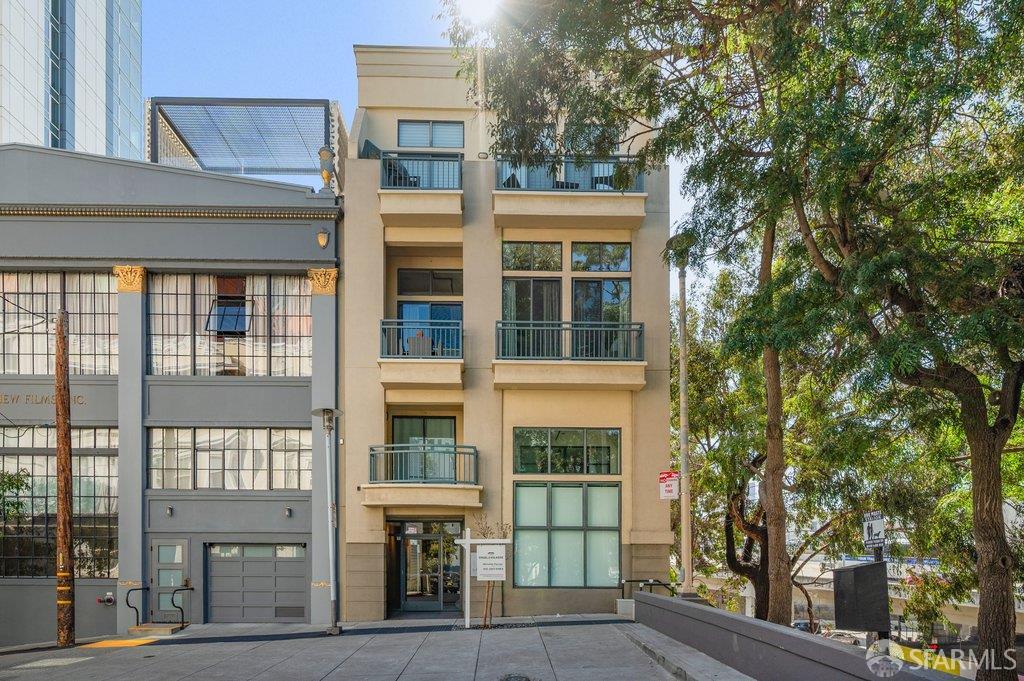 Beige multi-story building with balconies and large windows, framed by leafy trees.