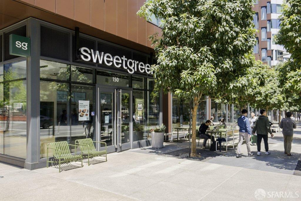 Sweetgreen storefront with sidewalk seating and leafy trees along a sunny city street.