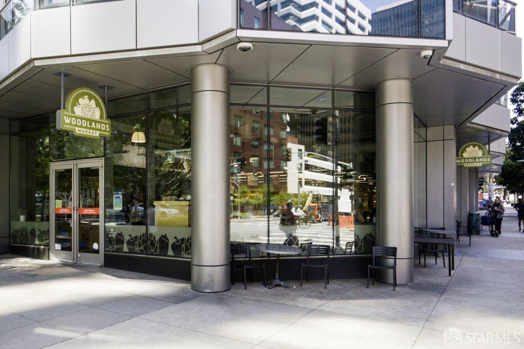 Woodlands Market storefront beneath a modern building, with metal columns and sidewalk seating.