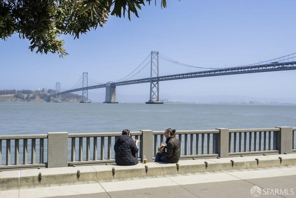 San Francisco Bay waterfront with the Bay Bridge and two people seated by the railing.