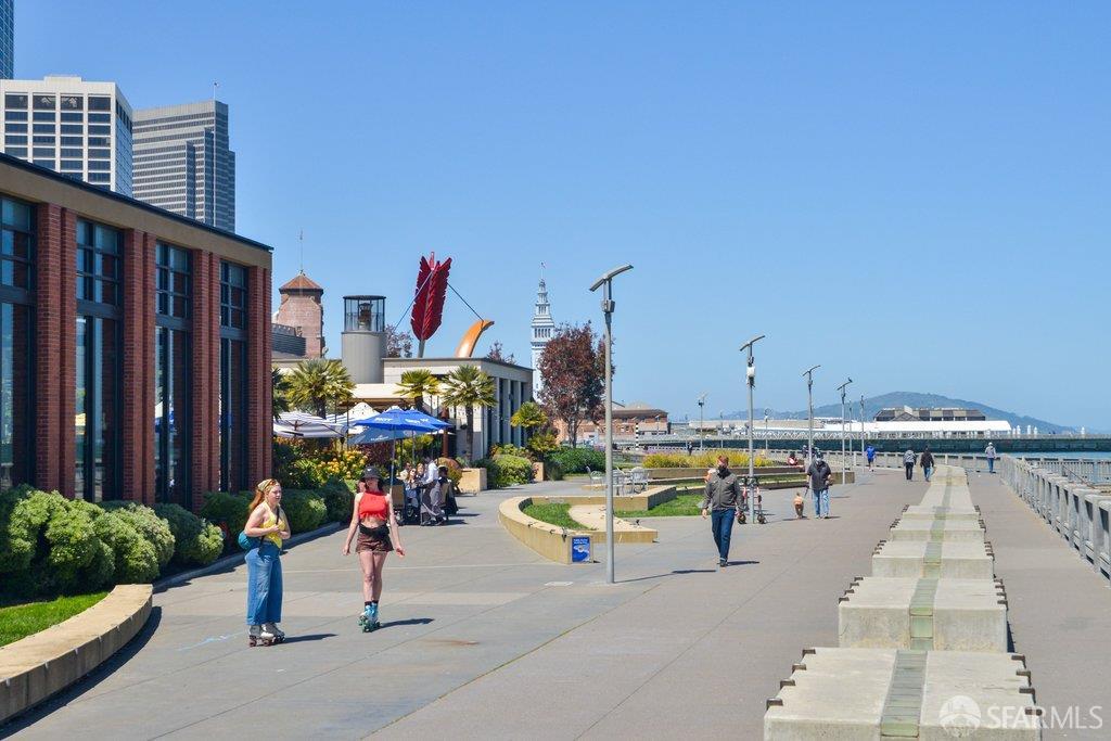 San Francisco waterfront promenade with pedestrians, landscaped planters, and city buildings.