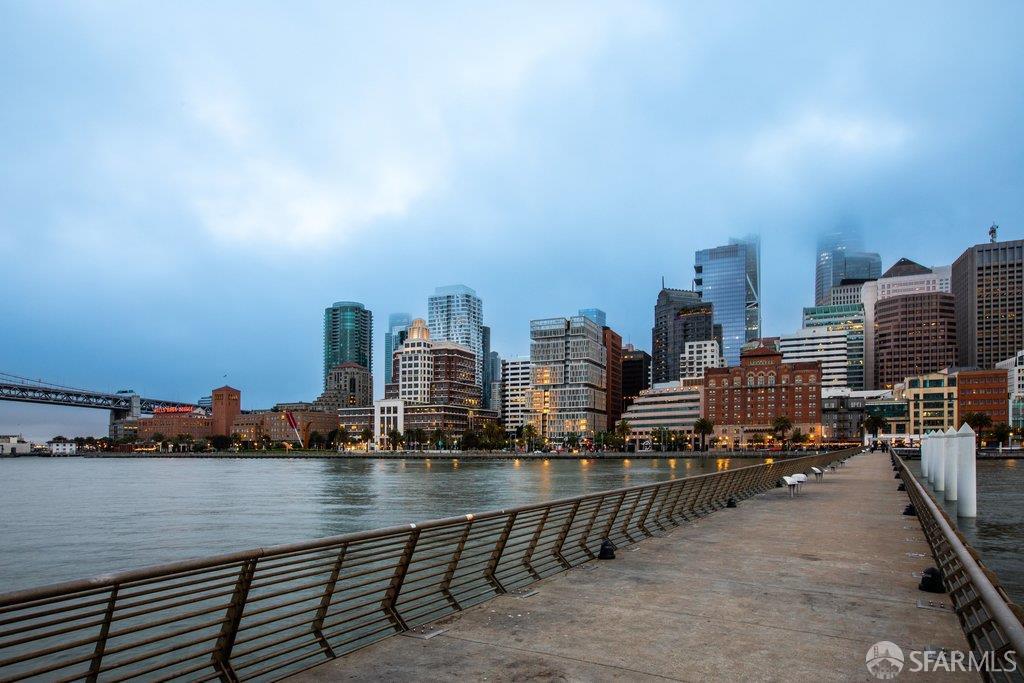 San Francisco waterfront promenade and skyline across the water, with a bridge at left.