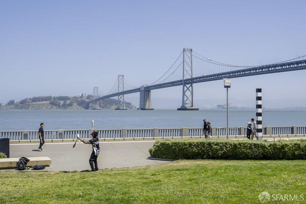 Bay Bridge across the water, viewed from a grassy waterfront promenade with pedestrians.
