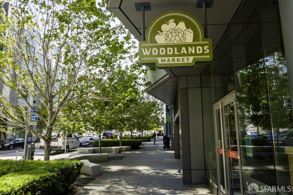 Woodland Market storefront with a green hanging sign beside a tree-lined sidewalk.