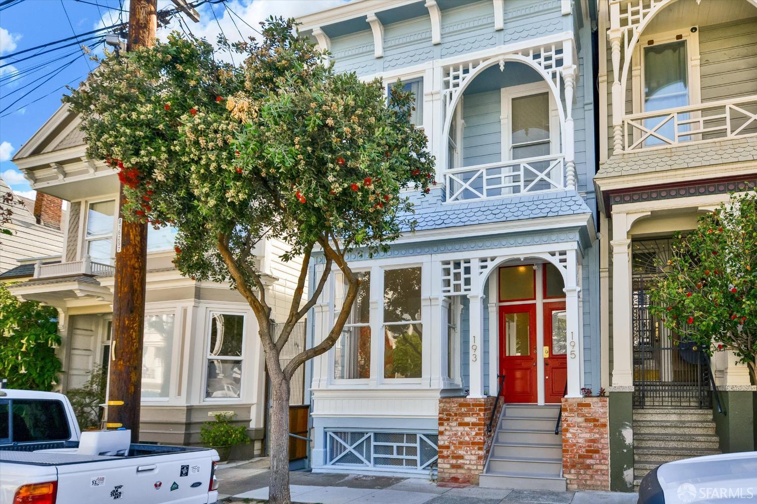 Blue Victorian home with ornate white trim, red double doors, and a leafy flowering street tree.