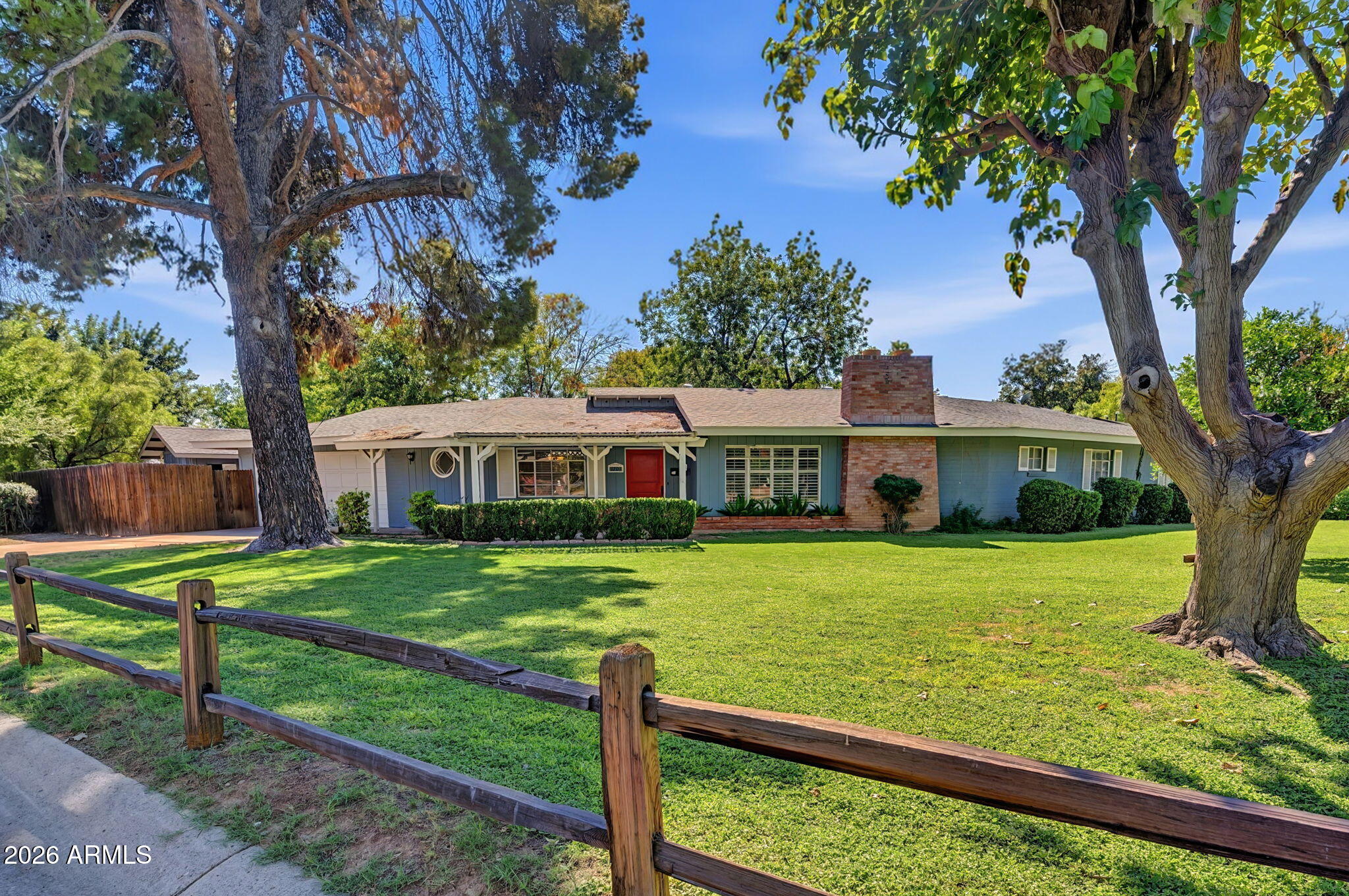 Blue single-story home with a red door, brick chimney, and mature trees framing the lawn.