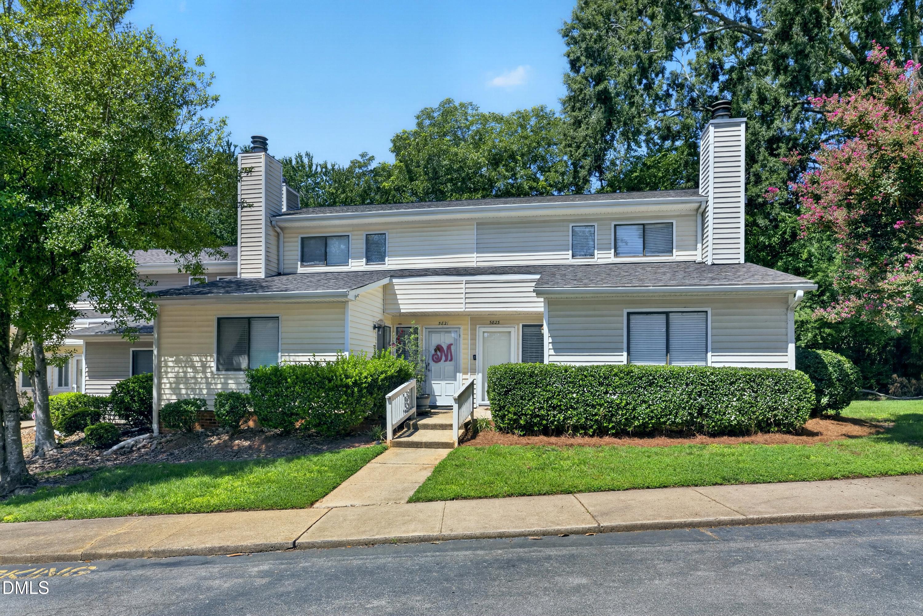 Two-story beige townhouse with twin chimneys, trimmed hedges, and a front walkway.