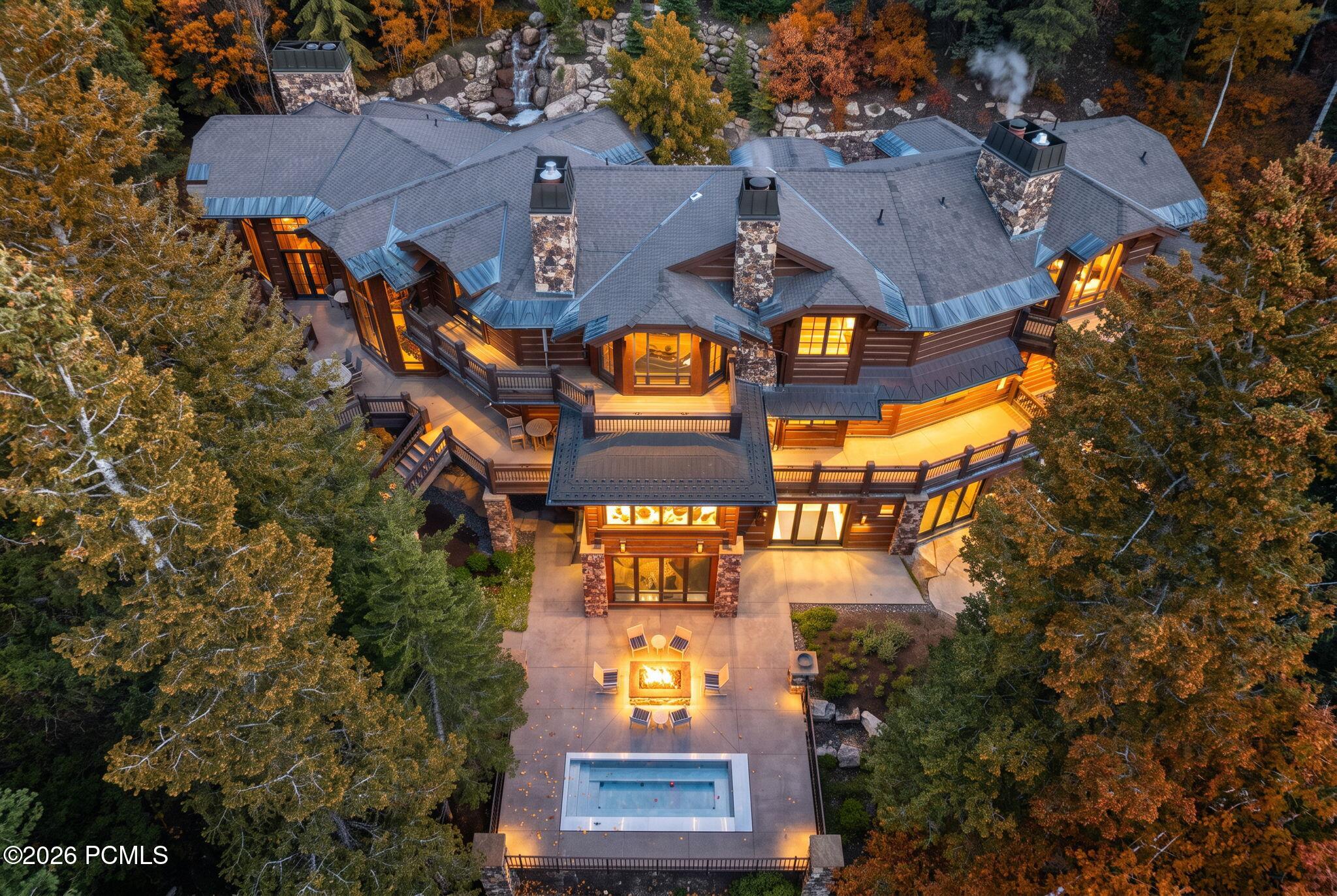 Aerial view of a timber-and-stone home, pool, and fire pit among autumn trees.