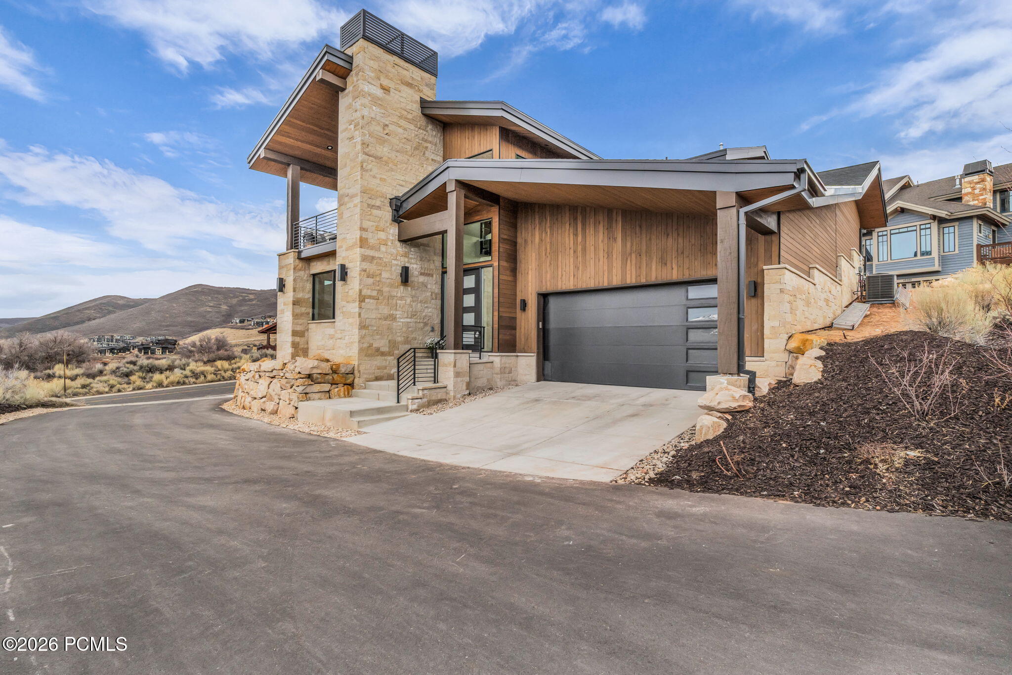Modern stone-and-wood mountain home with a tall chimney, wide garage, and dry hills behind it.