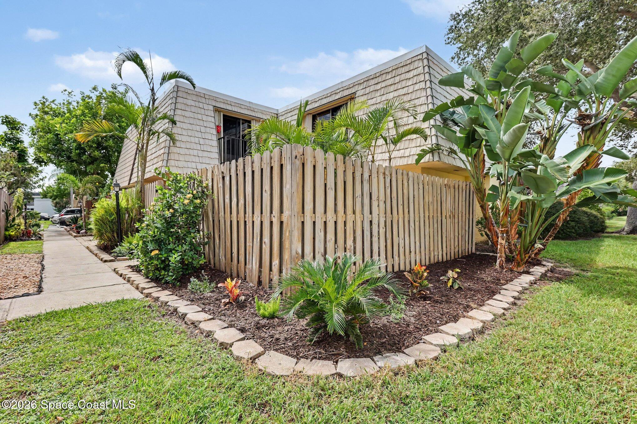 Two-story shingle-sided home behind a tall wooden privacy fence, surrounded by tropical landscaping.