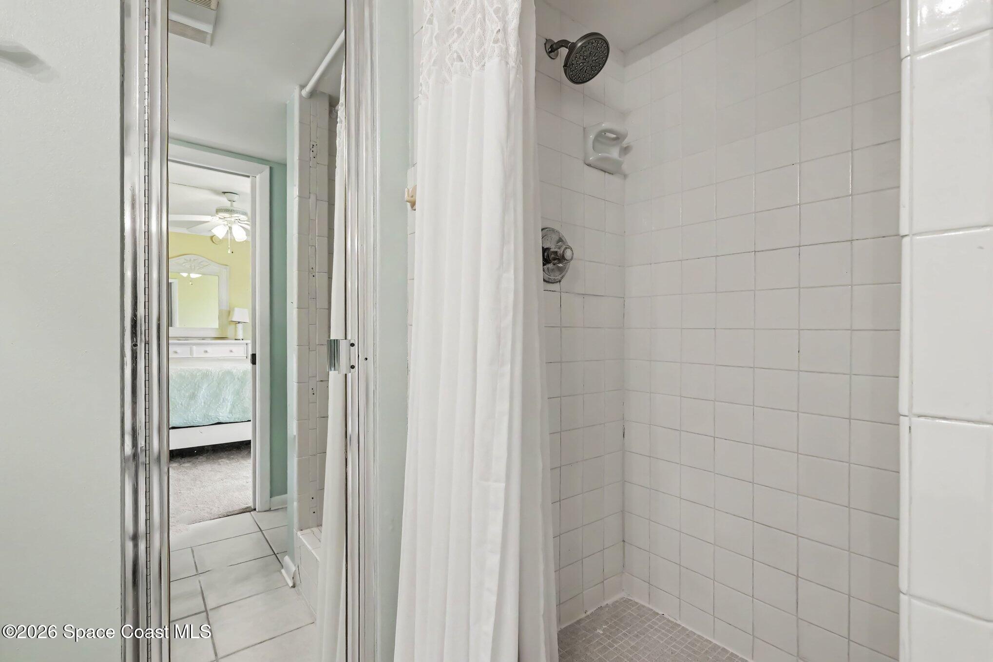 White-tiled shower with a dark showerhead and a view into the adjoining bedroom.