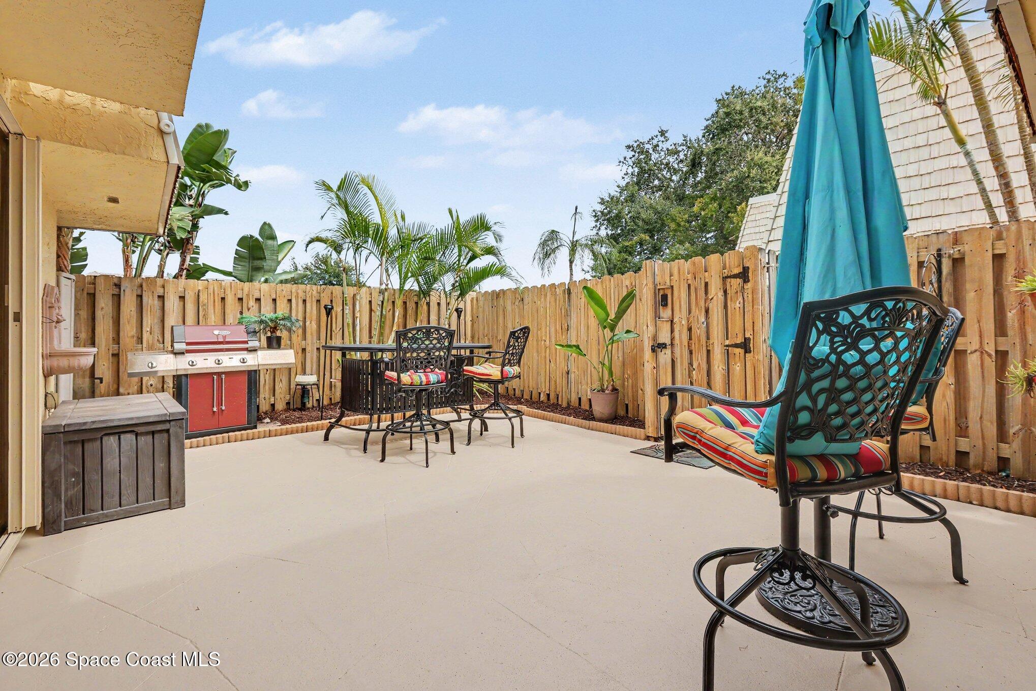 Fenced patio with a red grill, outdoor dining set, and metal lounge chairs beneath a turquoise umbrella.