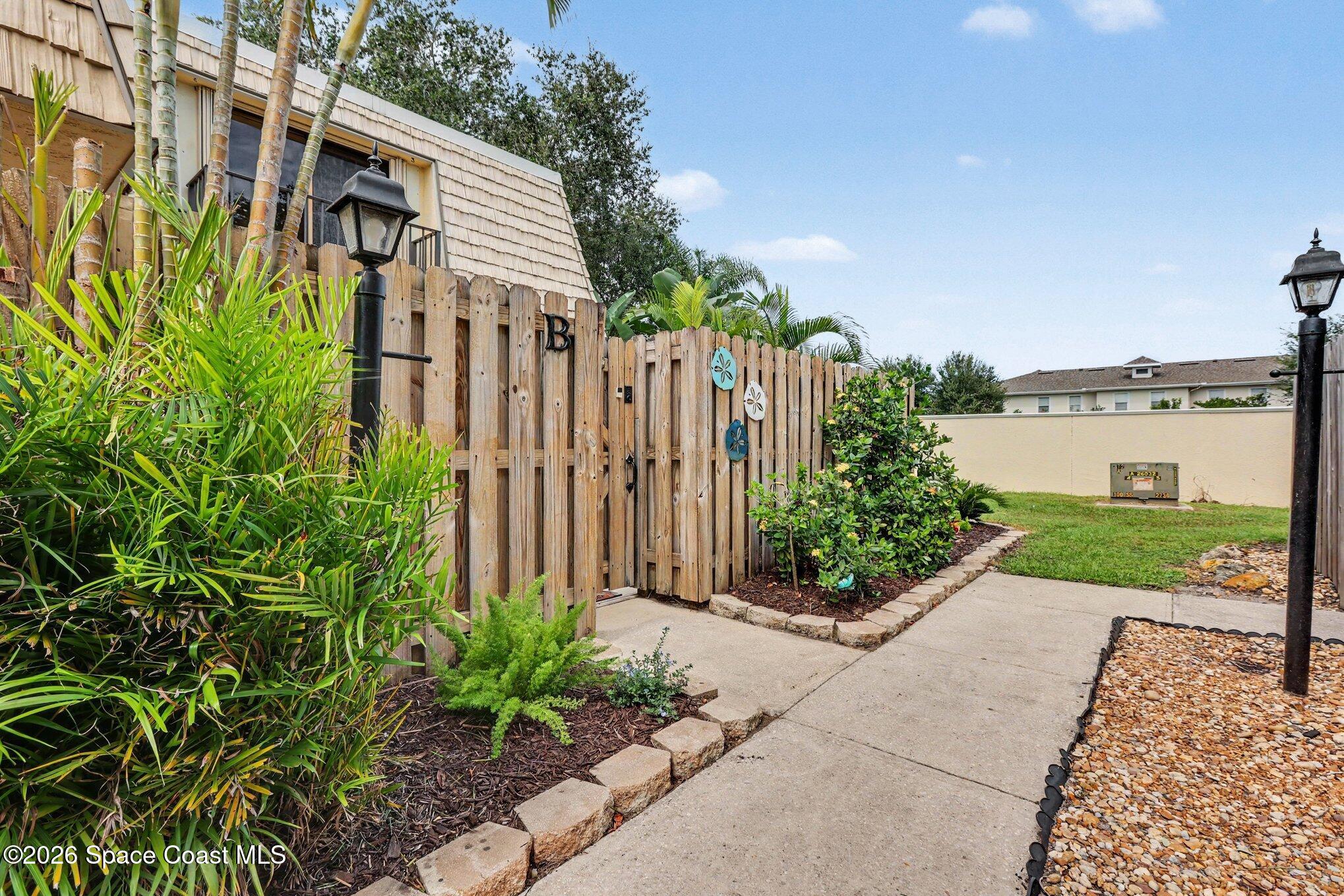 Residential entrance with a wooden gate marked B, tropical plants, and a paved walkway.