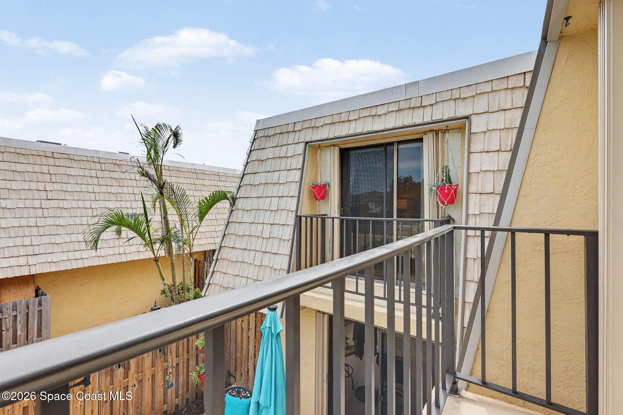 Condo balcony with dark metal railings, red hanging planters, and a view of neighboring shingle-sided buildings.