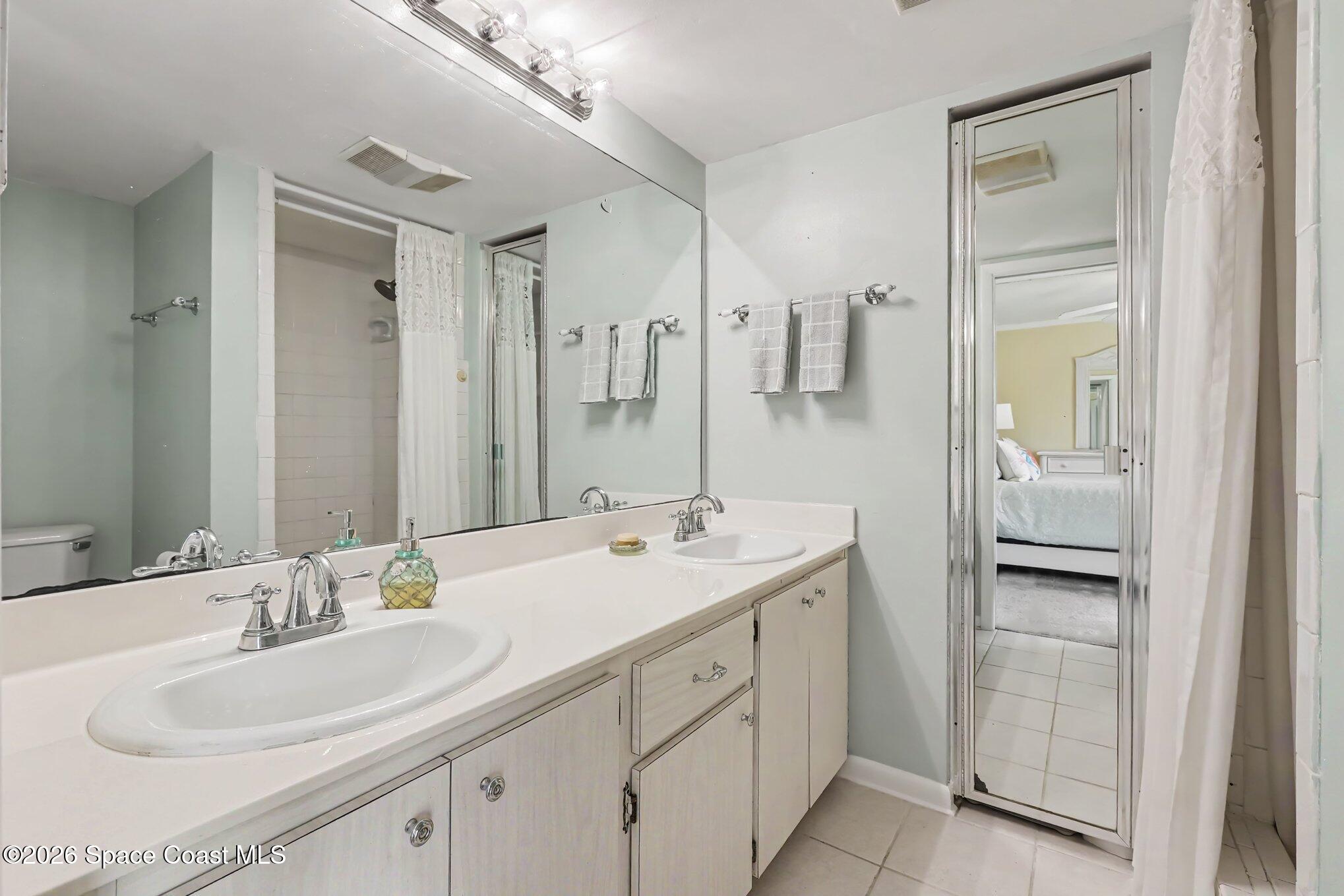 Bathroom with a long double-sink vanity and mirrored door reflecting an adjoining bedroom.