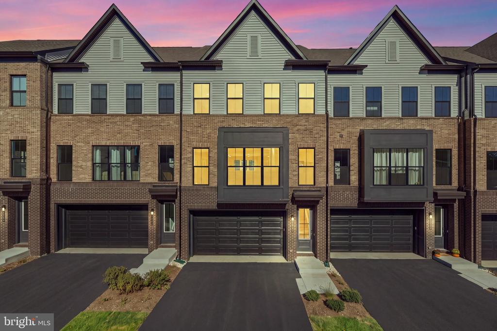 Attached brick townhouses with gray gables, front garages, and warmly lit windows at dusk.