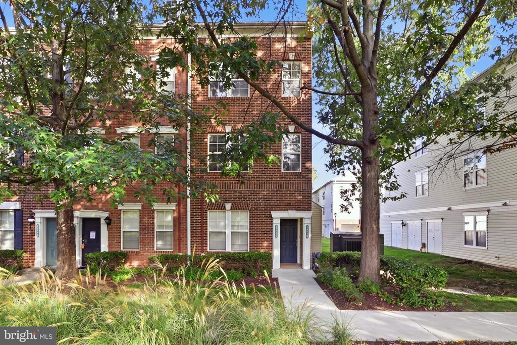 Brick townhome exterior with a front walkway, mature trees, and landscaped plantings.