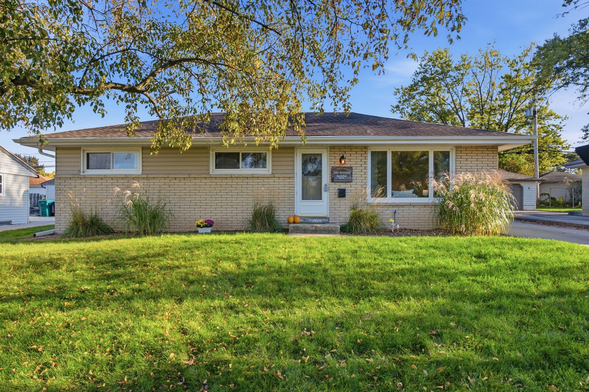 Single-story tan brick home with a broad lawn, mature trees, and ornamental grasses by the front entrance.