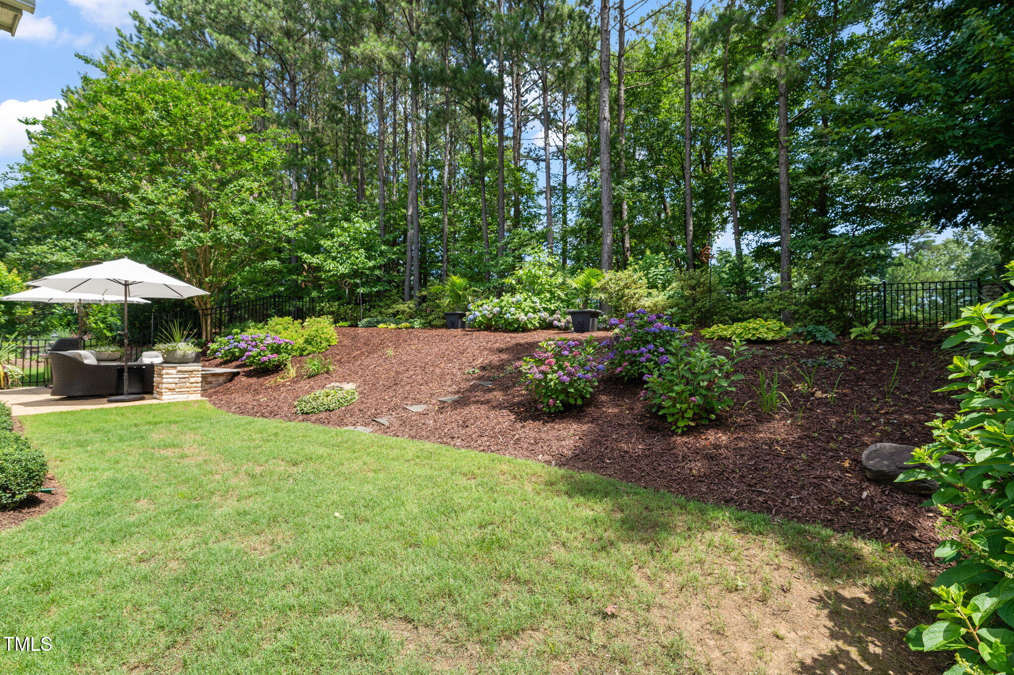 Landscaped backyard with flowering hydrangeas, mature trees, and a patio seating area beneath a white umbrella.