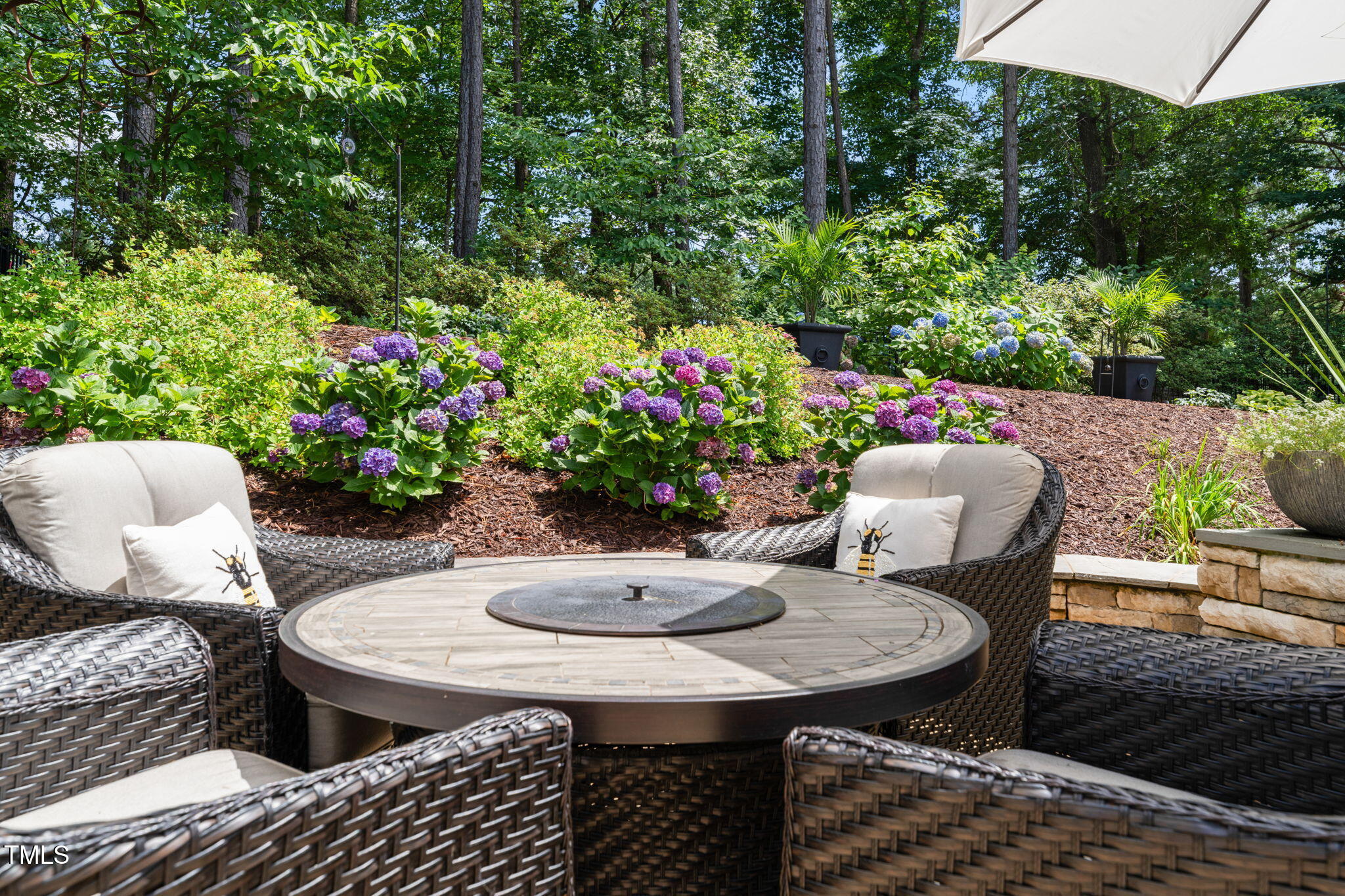 Furnished patio with wicker seating and a round fire table beside colorful hydrangeas.