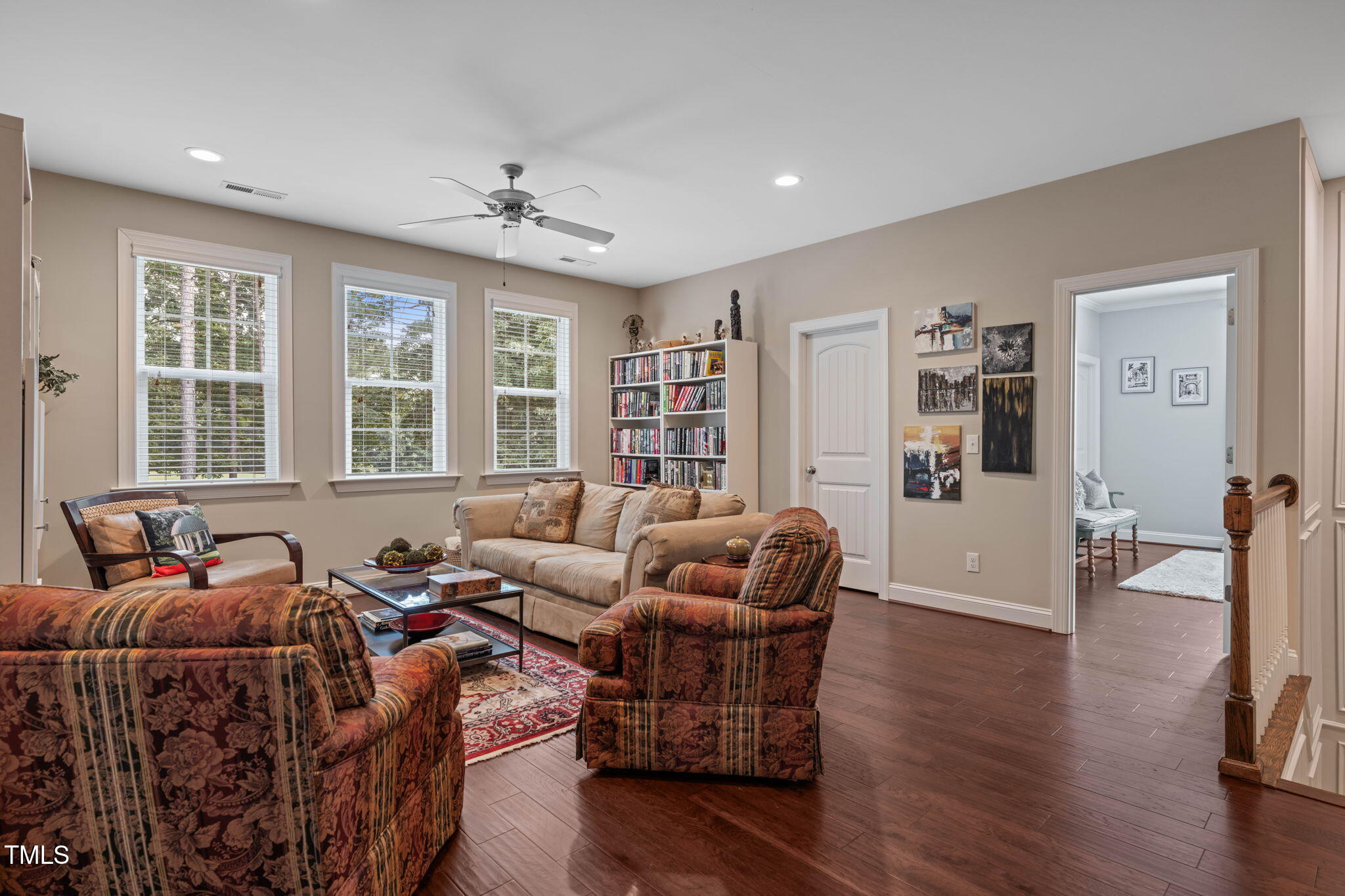 Living room with a bookcase, patterned armchairs, sofa, and three large windows.