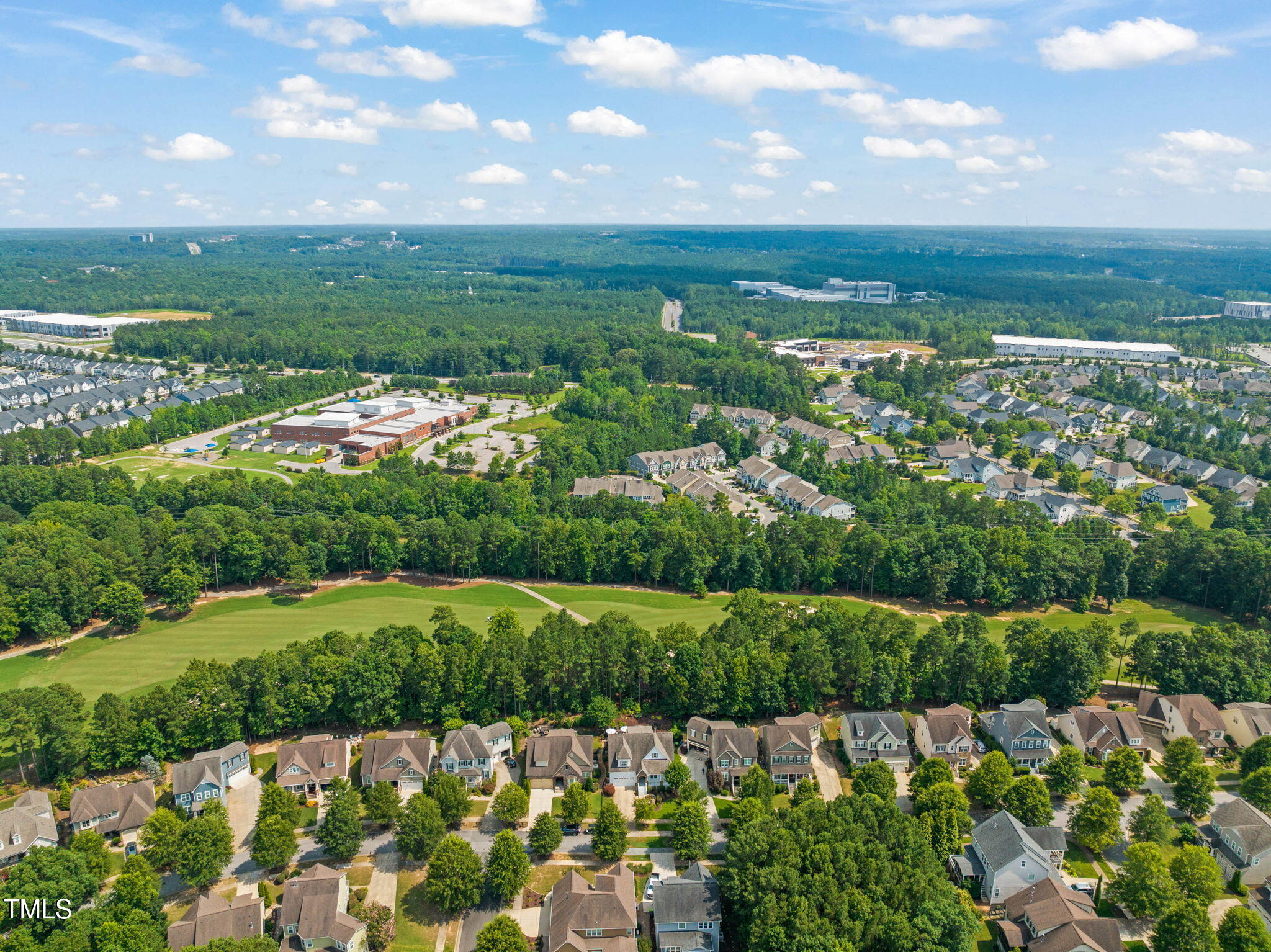 Aerial neighborhood view with a manicured green, dense trees, and homes stretching toward the horizon.