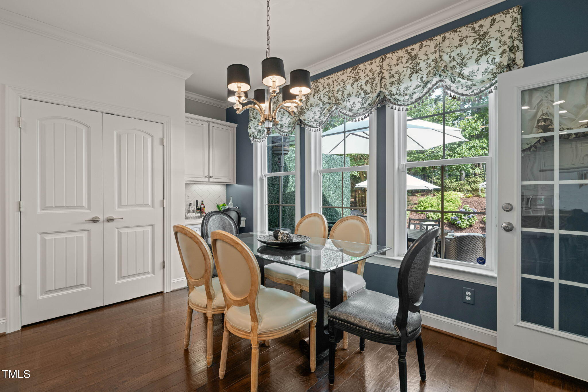 Dining room with a glass-top table, chandelier, and large windows overlooking a garden.