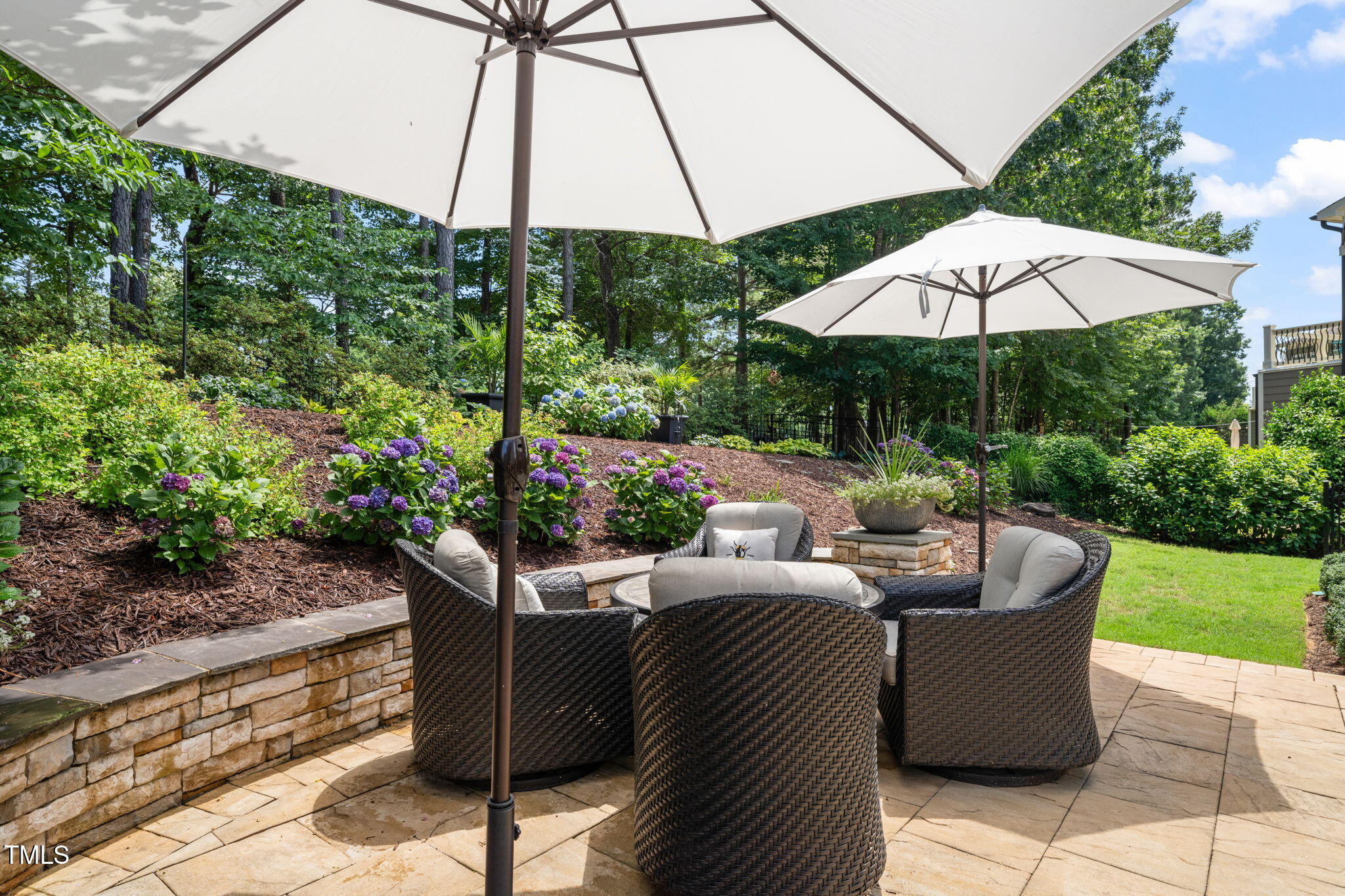 Furnished stone patio with wicker chairs and umbrellas beside a flowering garden.