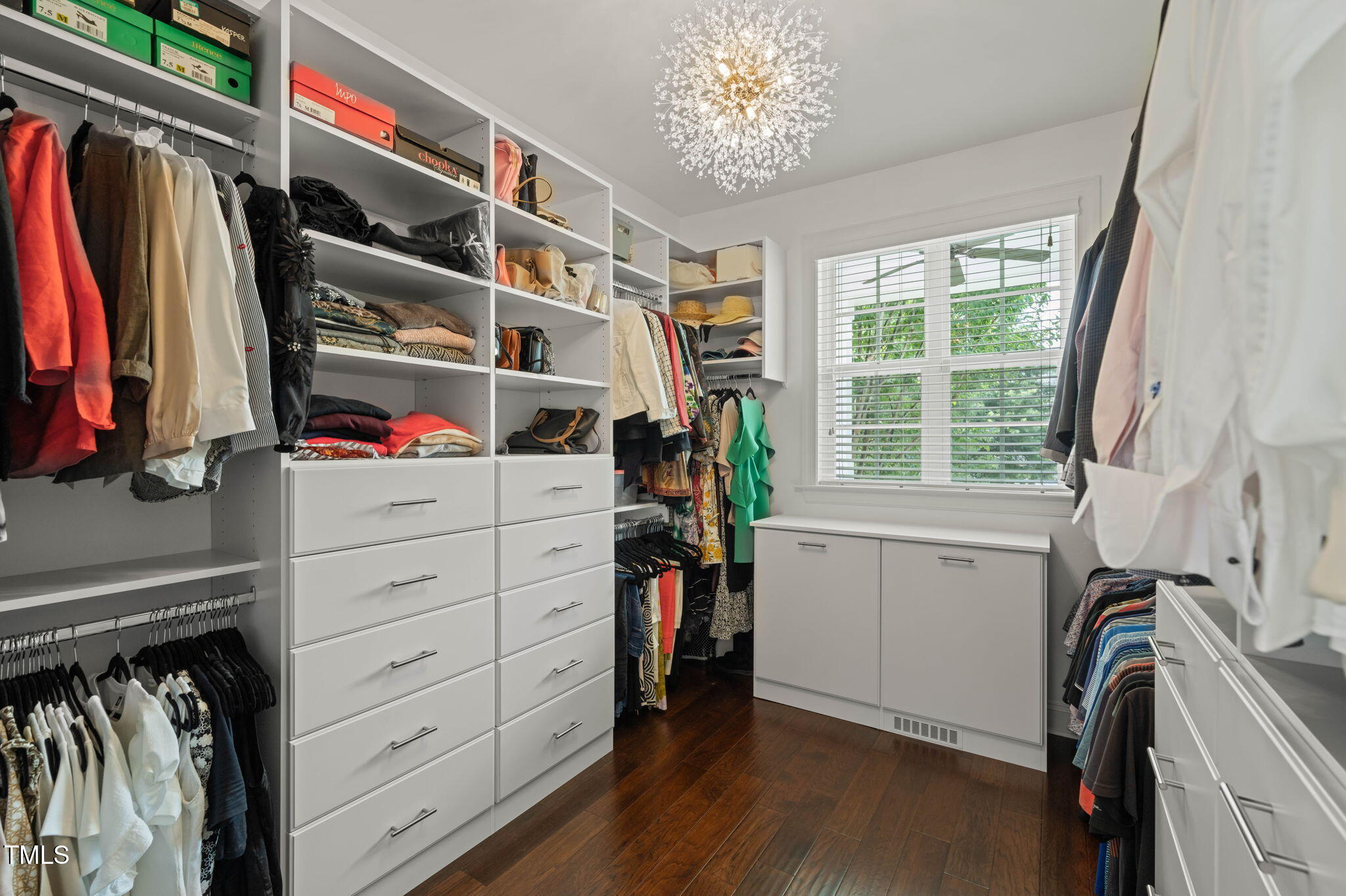 Walk-in closet with built-in white storage, hanging clothes, dark wood flooring, and a sparkling chandelier.
