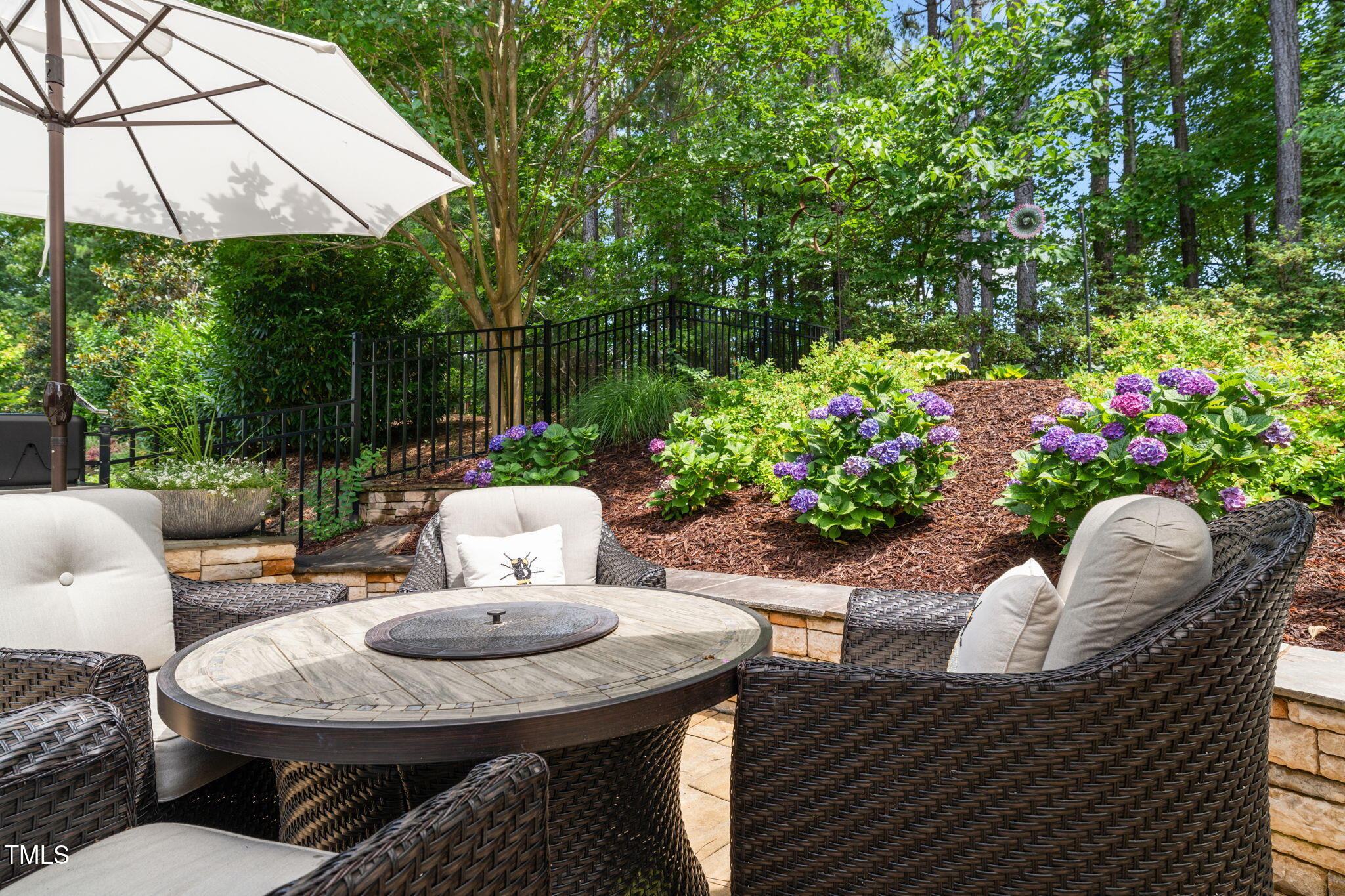 Outdoor patio with wicker seating, a large umbrella, and purple hydrangeas on a landscaped slope.