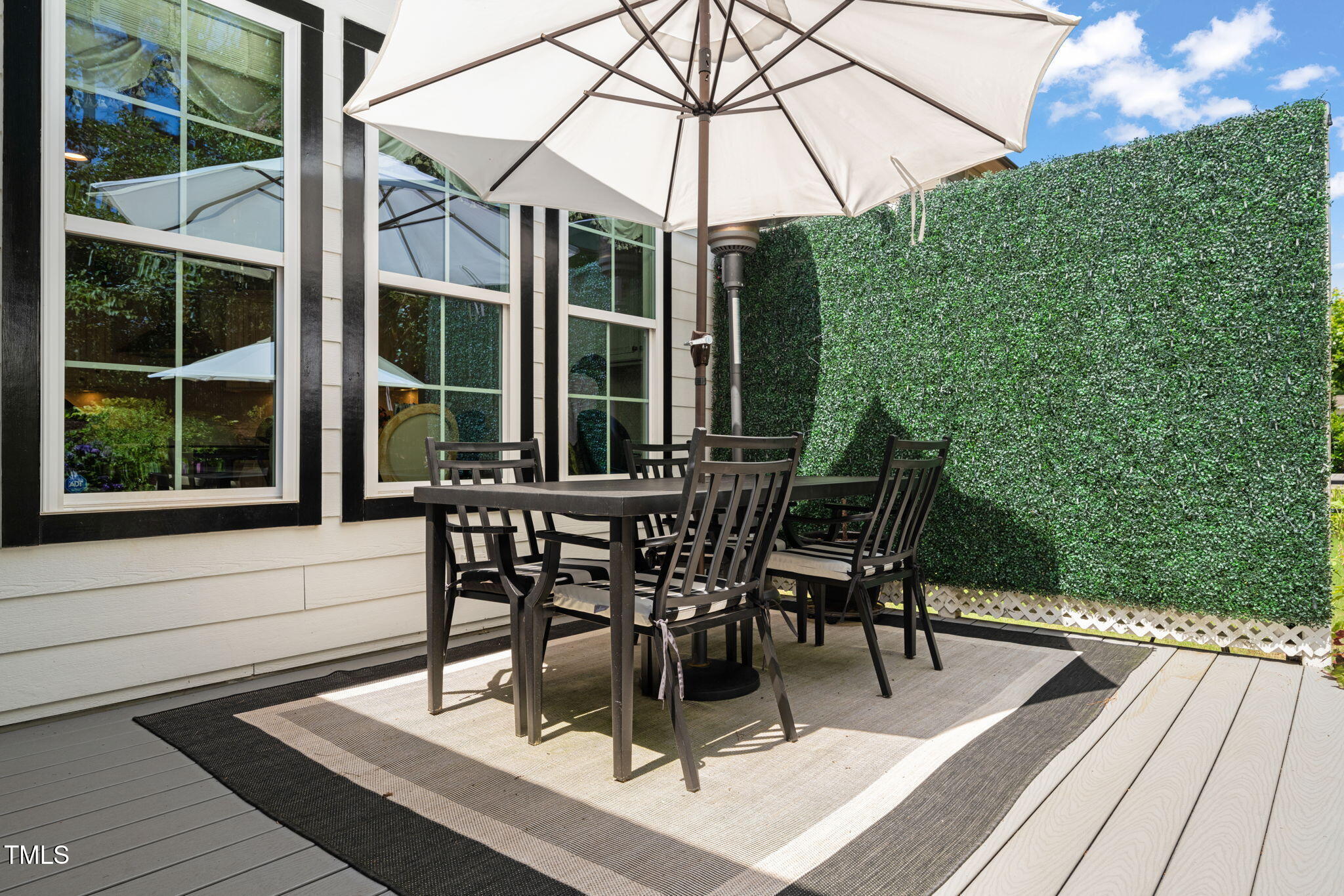 Outdoor dining set beneath a large umbrella on a deck beside a tall privacy hedge.