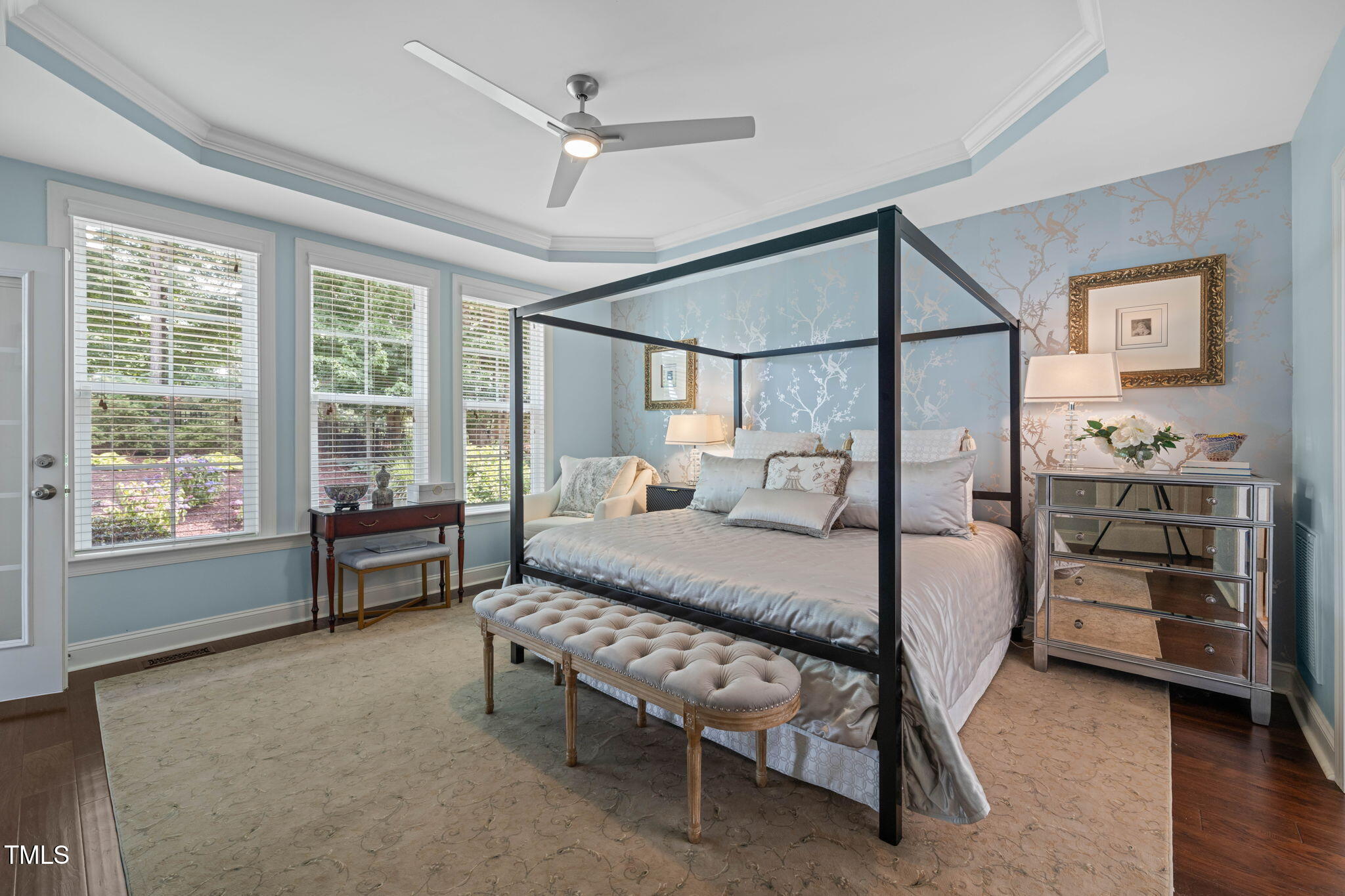 Furnished bedroom with a black four-poster bed, pale blue patterned wallpaper, and windows overlooking a garden.