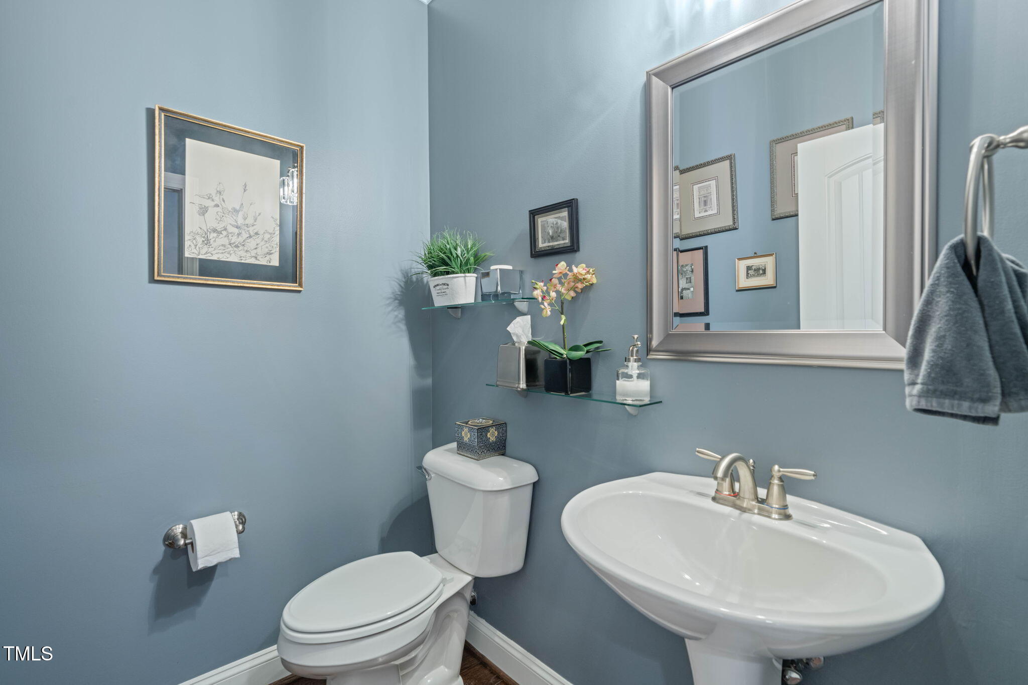 Blue-gray bathroom with a white pedestal sink, toilet, large mirror, and glass shelves.
