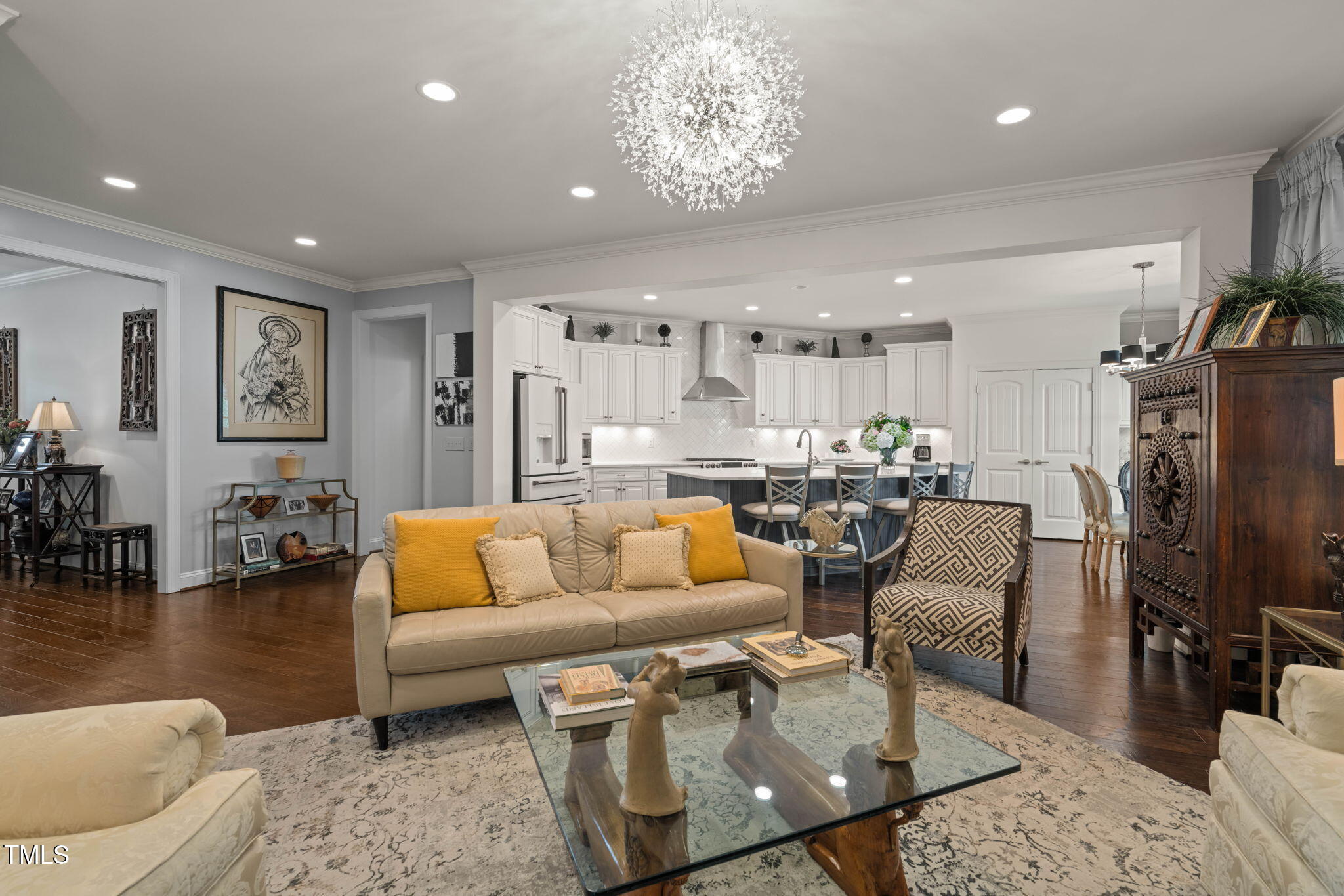 Open-plan living room and kitchen with a crystal chandelier, mustard pillows, and dark hardwood floors.