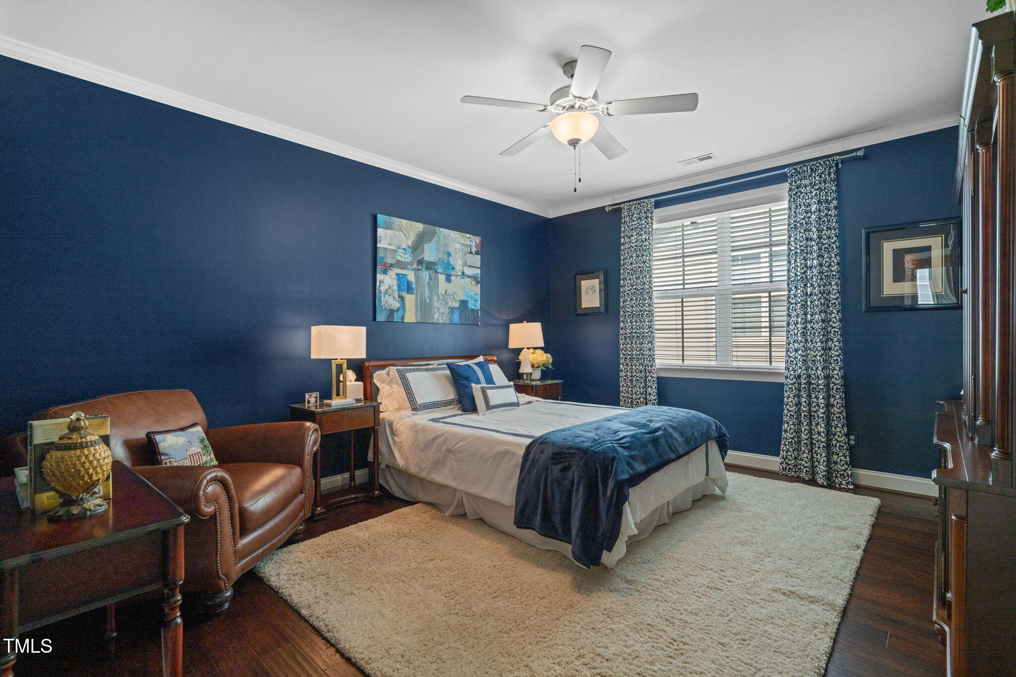 Furnished bedroom with dark blue walls, a white bed, navy throw, brown leather armchair, and window with patterned curtains.