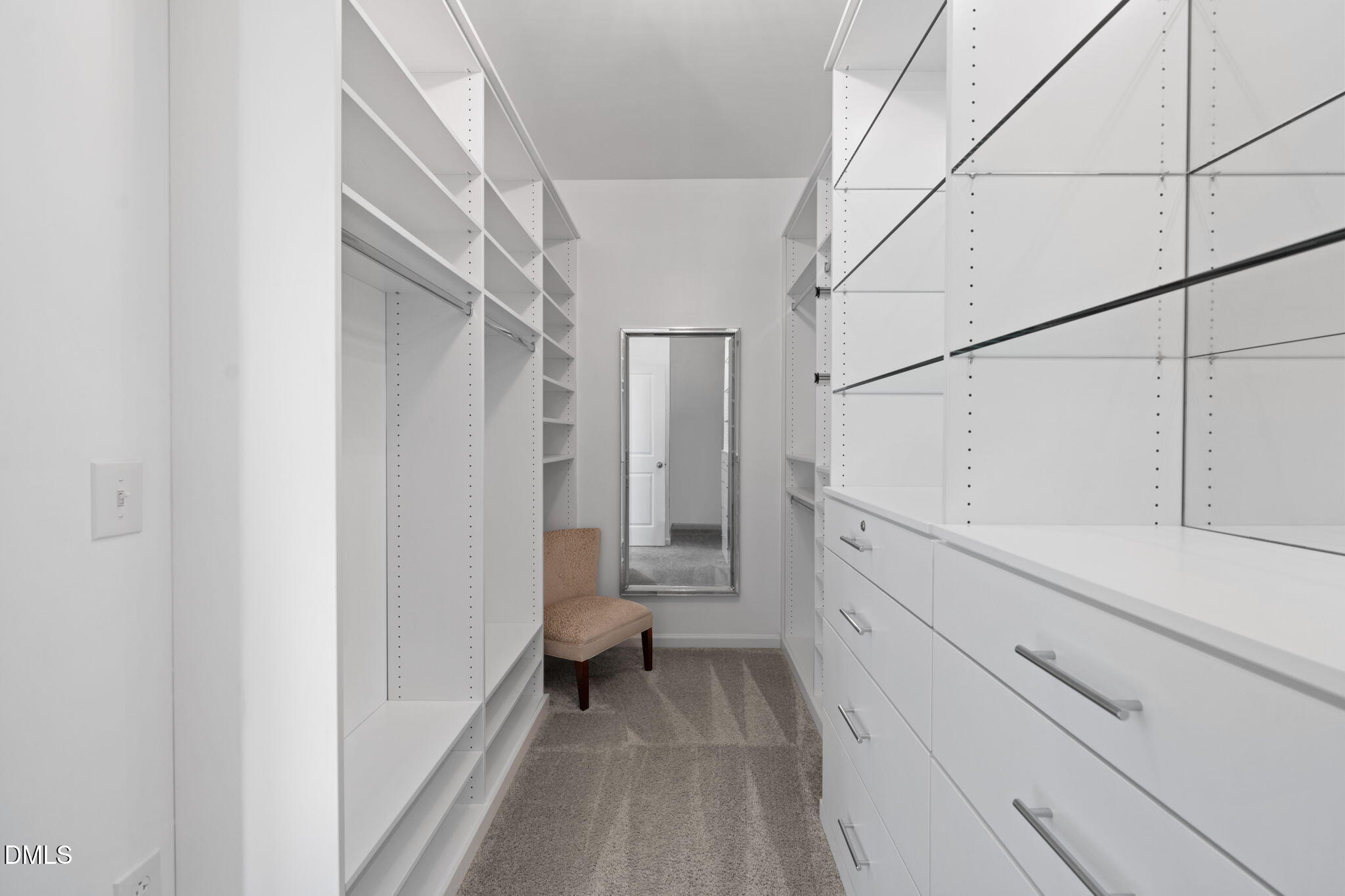 Walk-in closet with white built-in shelving, drawers, gray carpet, and a chair beside a full-length mirror.