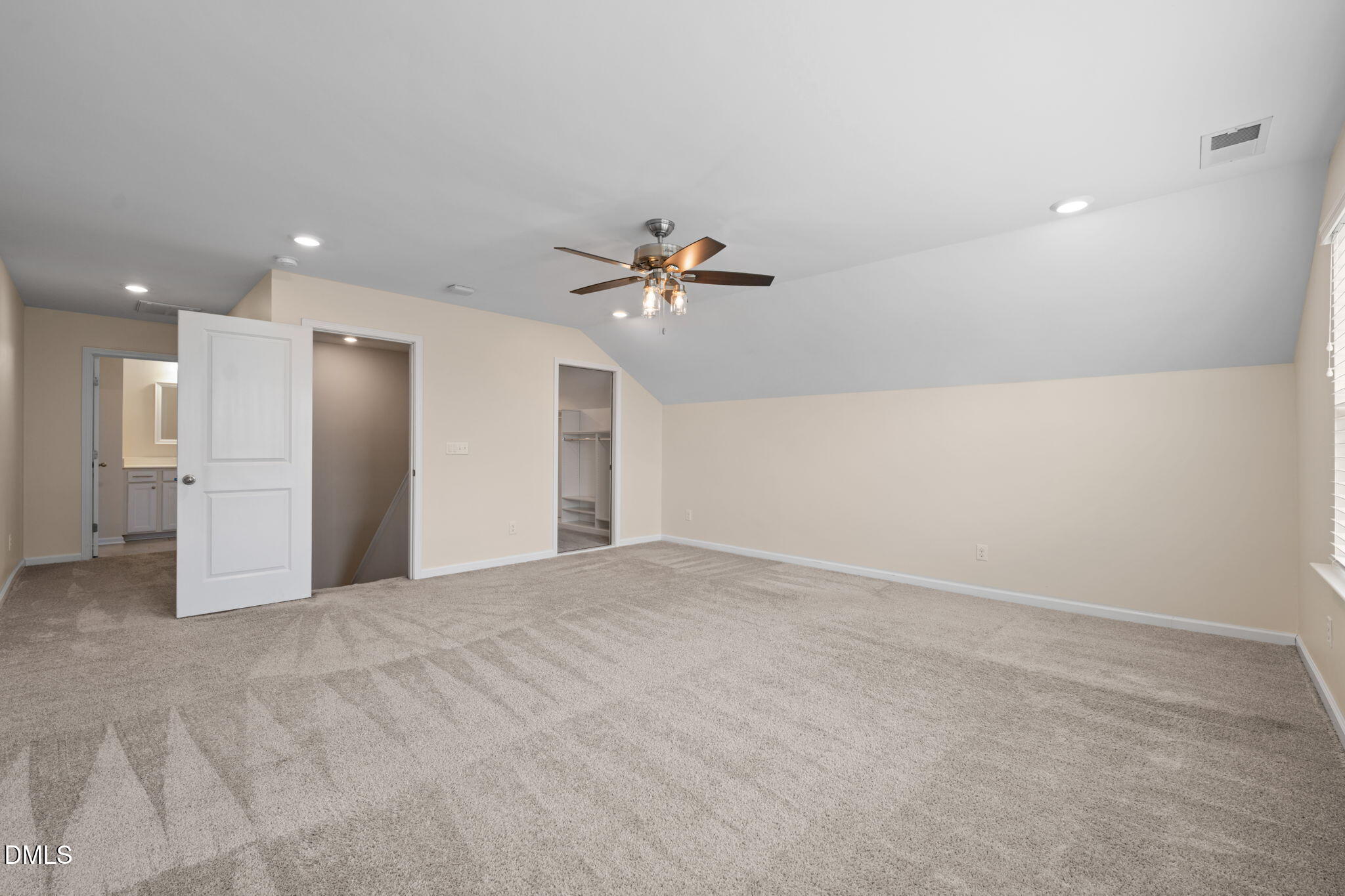 Empty carpeted bedroom with a sloped ceiling, ceiling fan, open closet, and bathroom vanity visible through the doorway.